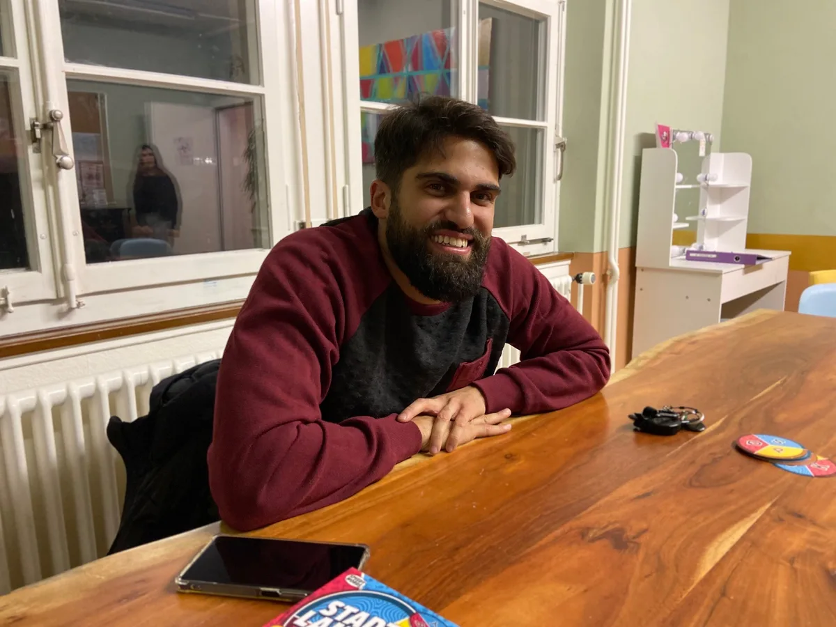 Shervin Aiyobi begleitet das Projekt «Jugendjobs» und vermittelt die Auftraggeber an die Teenager. (Archivfoto) Ein junger Mann sitzt an einem Holztisch und strahlt in die Kamera.